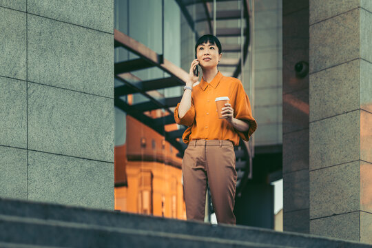 Young professional woman talking on phone while holding coffee outside modern office building