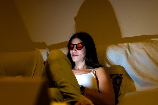 Woman relaxing on sofa, wearing orange tinted blue light blocking glasses at night