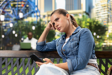 Fototapeta premium Thoughtful woman rests her head on one hand while holding a smartphone on a park bench, taking a reflective pause after a disappointing message.