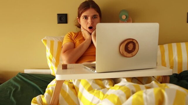 Shocked young woman looking at her laptop screen with an open mouth while sitting in bed under a yellow striped blanket