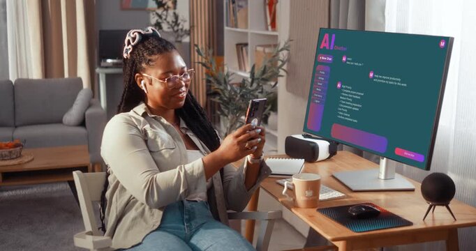 Smiling young Black woman relaxing and using her smartphone while sitting at the home desk in the living room, with an AI chatbot interface visible on the monitor of the computer.