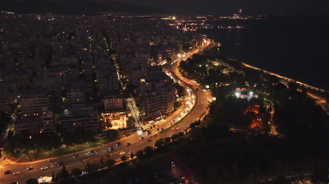 Night drone view of Poseidonos Avenue, Athens. The illuminated coastal road curves along the Aegean Sea, with the urban sprawl glowing under a bright full moon.