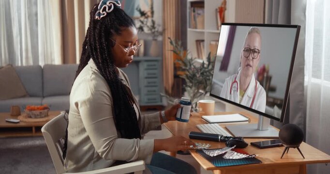 Young Black woman holds pills and consults a female doctor during a video call on computer from home, showing telemedicine, remote healthcare and medical advice.