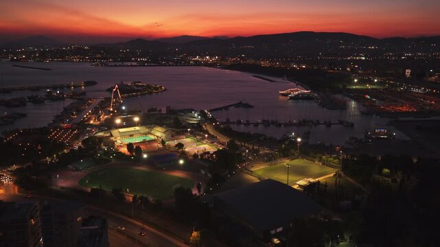 Amazing aerial perspective of a coastal city at twilight, showing the harbor, marina, and illuminated sports facilities as the vibrant orange sky transitions into a deep night blue