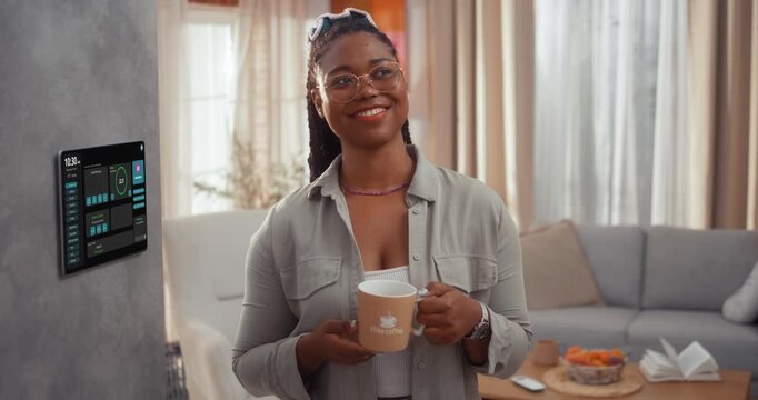 Smiling young Black woman activates an AI home assistant on a wall smart home touchscreen and looks at camera while holding a coffee cup in a modern living room.