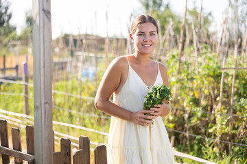 Obraz premium European girl stopped near bed of tomatoes, utility shed and barn, takes break from gardening, enjoys active hobby of growing garden crops. Girl with agricultural tool in hands, stands near trellis