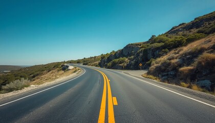 Obraz premium Asphalt highway curving through rugged coastal terrain under clear blue summer sky during bright daylight with hilly landscape under bright depicts empty asphalt