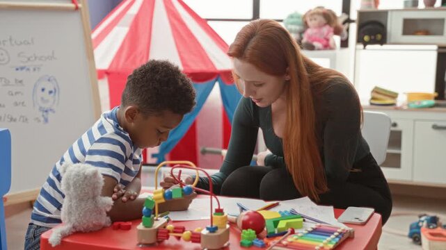 Woman guiding boy drawing and writing at a toy covered table in a building playroom with a red tent and whiteboard; learning concentration.