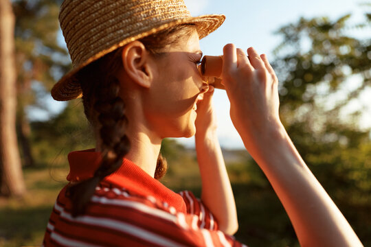 A young girl in a striped shirt and straw hat peers through binoculars in a sunlit park, capturing curiosity, exploration, and a moment of private discovery as birds sing and a gentle breeze rustles