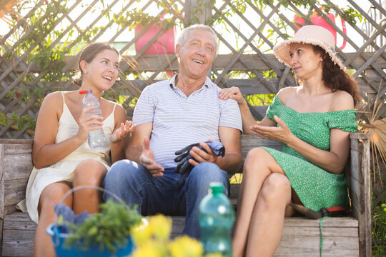 Elderly man and two women are chatting cheerfully while sitting on bench in the courtyard of a country house