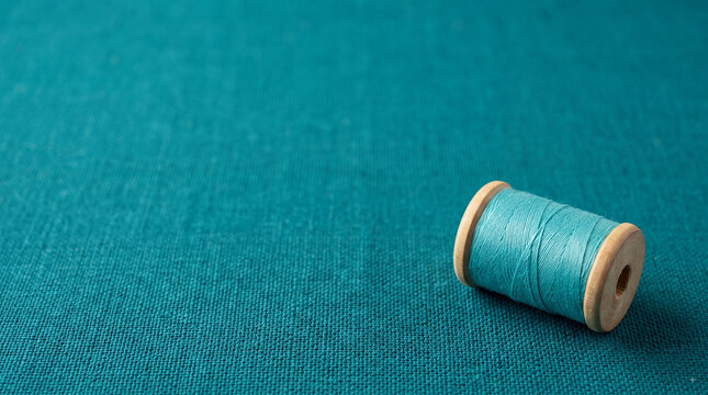 Teal sewing thread spool on matching textured fabric with soft light, minimal craft background and copy space for tailoring or textile design