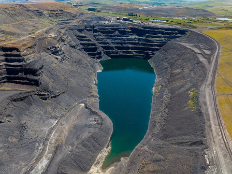 Aerial view of closed open pit coal excavation site, flooded void surrounded by dark terraced earth and access tracks, environmental impact of fossil fuel extraction