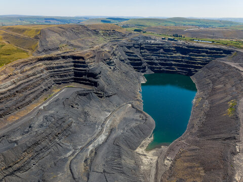 Deep opencast coal mine pit filled with green water, former extraction benches and terraced earthworks exposed under bright daylight in South Wales