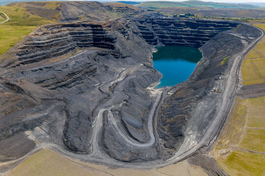 Abandoned opencast coal mine pit with deep blue water flooded at bottom, steep terraced excavation levels and tracks cutting through dark earth, high angle view