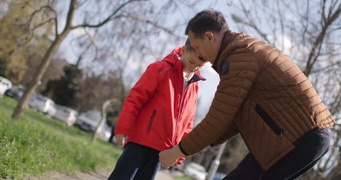 Father assists his son with putting on a jacket as they enjoy a sunny day in a park. They walk along the paved path surrounded by trees and other families