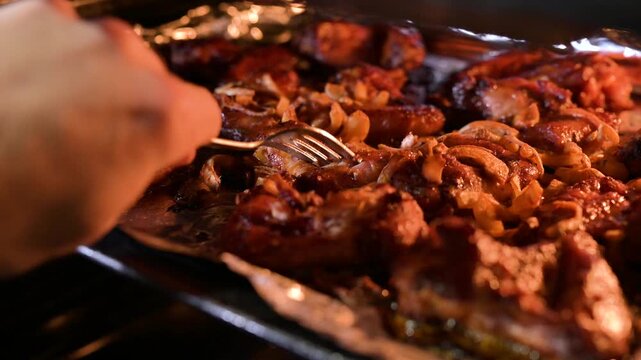 A man checking the readiness of baked chicken wings in the oven. Male hand pierces roasted meat with onions on a baking sheet, reflecting culinary skill and satiety