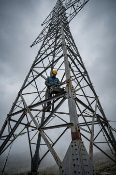 Electrician working on high voltage pylon, low angle shot, safety helmet and tools, suitable for energy industry, infrastructure maintenance and power supply advertising
