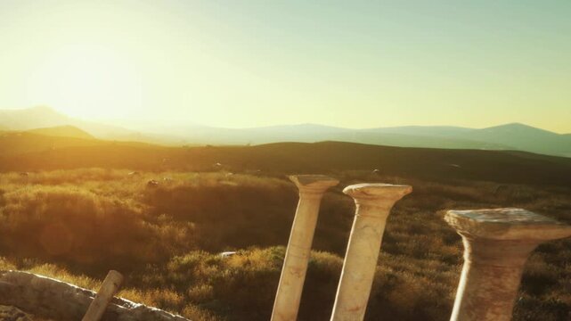 Four old pillars are seen against a sunset in the countryside. The landscape is wide and open, with distant mountains visible. The scene shows a quiet moment in history.