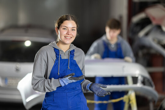 Service station young woman employee works in repair box room, department. Worker performs car body repairs and installs spare parts on vehicle body.