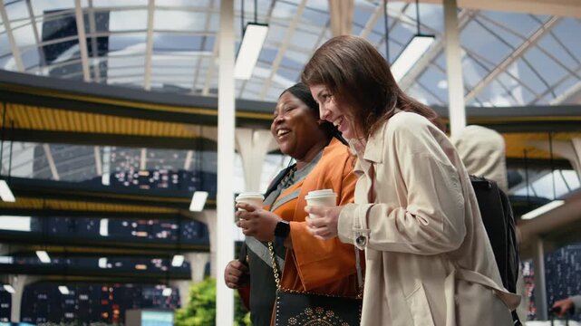 Friends carry drink cups through a coach station while discussing travel plans; the companions enjoy refreshments and conversation before departure.