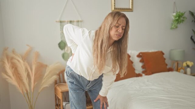 Woman holds lower back, bends toward bed with hand on knee and wincing in a bedroom inside a building; chronic pain.