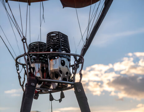A close-up of a hot air balloon burner assembly featuring two metal combustion heads mounted within a circular steel frame, suspended by cables beneath the balloon fabric against a cloudy sky.