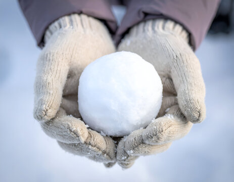 Person wearing beige knit gloves holding a compact snowball in their cupped hands against a snowy surface. Useful for winter, outdoor, cold weather, and play.