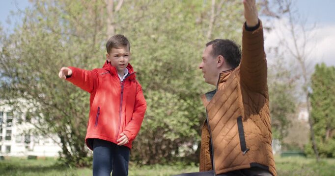 A father and his young son spend a sunny afternoon in a vibrant park. They are engaged in playful activities, surrounded by greenery and blooming trees, enjoying the warmth of spring