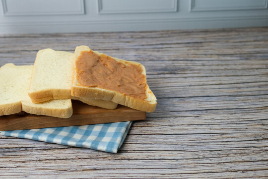 Soft bread slices rest on a checkered cloth. Wooden table shows weathered, natural grain. One slice topped with ritz, peanut jam. Simple, cozy, inviting breakfast scene. Ideal for morning meal, cafe