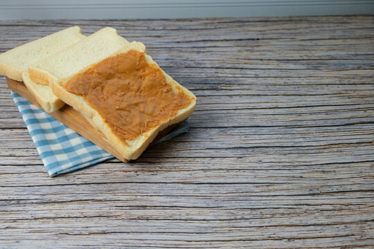 Soft bread slices rest on a checkered cloth. Wooden table shows weathered, natural grain. One slice topped with ritz, peanut jam. Simple, cozy, inviting breakfast scene. Ideal for morning meal, cafe