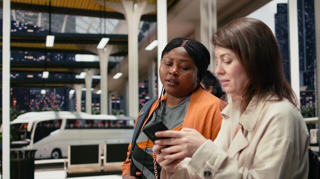 Friends review route details on a mobile phone while waiting in a coach station; the passengers walk together through the bus terminal and use a smartphone to manage their trip itinerary.