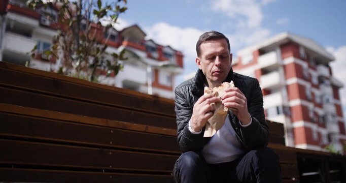 A man sits at a wooden bench in a lively urban park, savoring a tasty meal wrapped in paper. Sunlight illuminates the surroundings, creating a pleasant atmosphere for dining