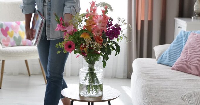 Woman taking magazines from sofa and bouquet of beautiful summer flowers indoors, closeup