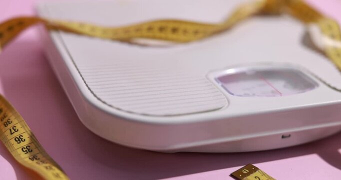 Scales with measuring tape on pink background, closeup. Camera moving left