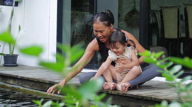 Happy Asian grandmother and her little granddaughter sitting on a wooden deck by a fish pond, smiling and enjoying a peaceful moment together in a backyard garden.