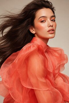 Confident young woman in voluminous coral pink ruffled blouse with dramatic wind-blown hair, glamorous portrait with soft studio lighting, high-fashion editorial concept for beauty and lifestyle