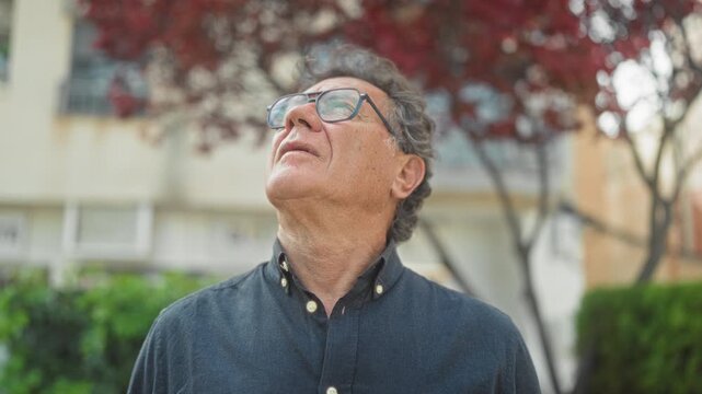 Middle aged man wearing glasses and a dark button shirt tilting his head back and looking up at red leaves by a building on a street; quiet contemplation.
