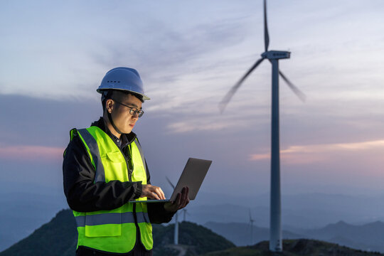 Engineer operating laptop on-site at wind farm during twilight with turbine in background