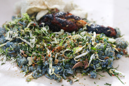 A fully mixed serving of Malaysian Nasi Kerabu featuring fragrant blue pea flower rice, grilled chicken, raw vegetable and herb salad (ulam), and crispy fish crackers.