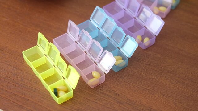 Colorful pill organizers in yellow, pink, and blue arranged on a wooden surface, showcasing a sequence of compartments filled with yellow capsules for daily medication management