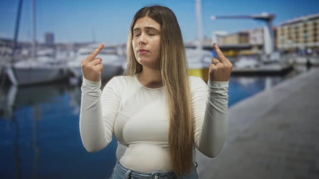 Woman showing both middle fingers on a street pier with boats, long hair and white long sleeve top, confrontational stare; defiance rebellion confrontation assertiveness.