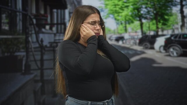 Woman pressing cheeks with hands, wearing glasses and jeans, pained expression on street beside building steps and parked cars; anxiety concern.