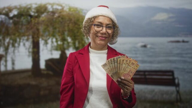 Woman holding a fan of banknotes with hand visible at a lakeside park bench, wearing santa hat and red jacket; holiday generosity cheerful.