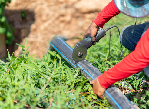 A worker using a handheld angle grinder to cut a black HDPE pipe outdoors on a grassy construction site.