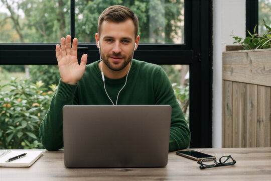Man in green sweater waves during video call while working remotely on laptop by window.