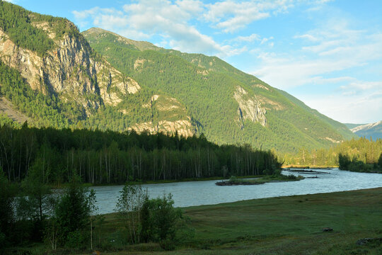 The winding bed of a wide river in the shadow of a mountain flows along mountain ridges illuminated by the rising summer sun in the early morning.