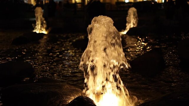Cinematic night fountain illuminated with warm golden lights in an urban park. Crystal-clear water streams rise and splash over dark stones, creating sparkling reflections and a peaceful evening 