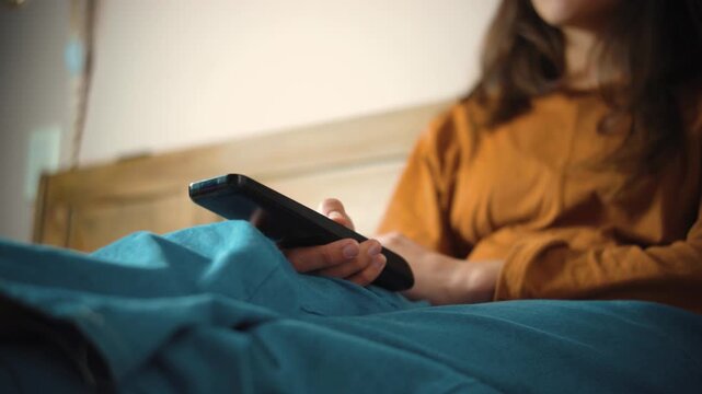 Young anonymous woman sitting on her bed while changing TV channels with a remote control, representing home entertainment, relaxation, leisure, and a casual moment at home.