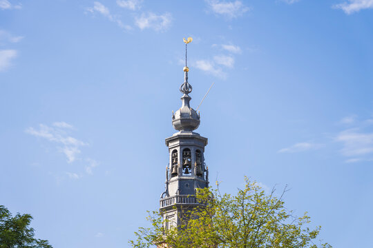 Low angle view of a historic church tower spire with bronze bells and golden weather vane against a cloudy blue sky backdrop