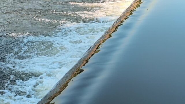 Fast-flowing water rushing over a small dam structure, creating white foam, turbulent currents, and dynamic cascades that highlight the power and movement of flowing water